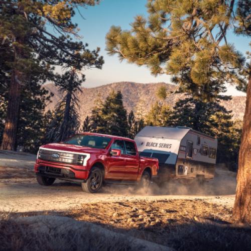 A red F-150 Lightning tows a camper in a forest. A mountain range rises in the background against a blue sky.