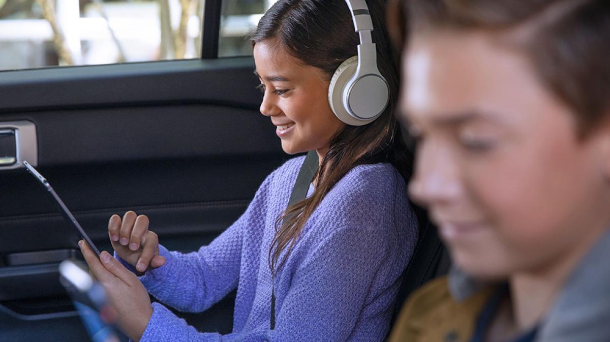 Two children smile while using a tablet and a phone in the back seat of a Ford vehicle.