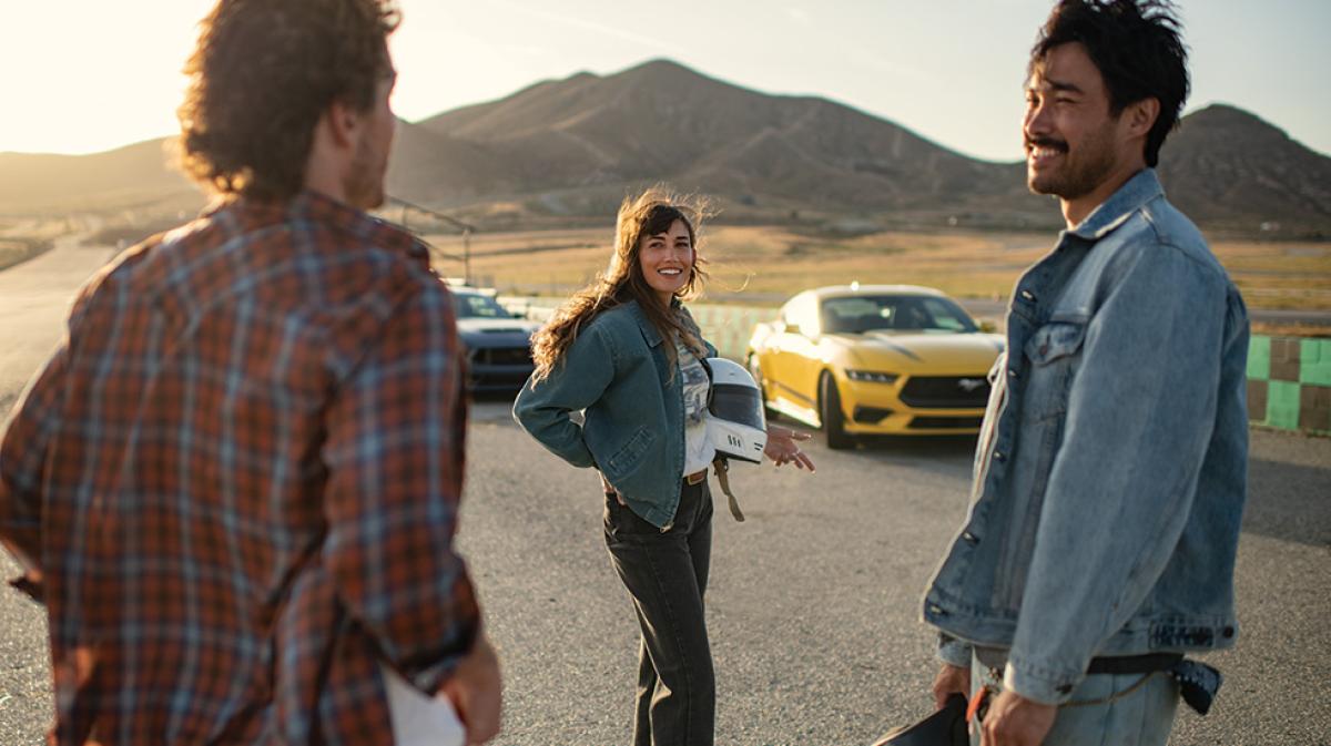 Friends are gathered at a closed track at magic hour, helmets in hands, with their Mustang coupes in the background
