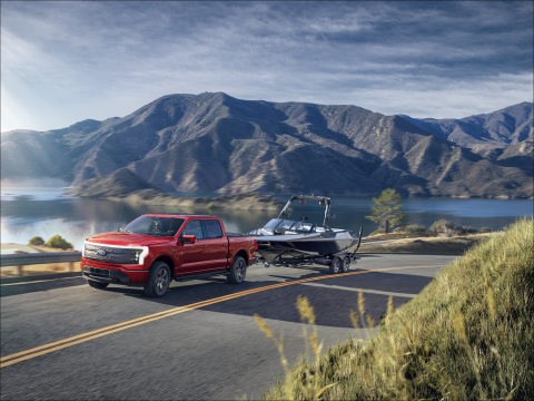 A red F-150 Lightning driving over an arching highway tows a boat. Mountains and a lake are in the background.